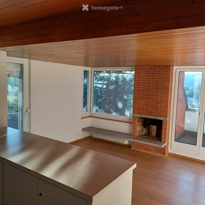 Empty room with wooden flooring, white walls, and a brick fireplace with a wooden bench. Large windows and sliding glass doors provide natural light and access to the outdoors.