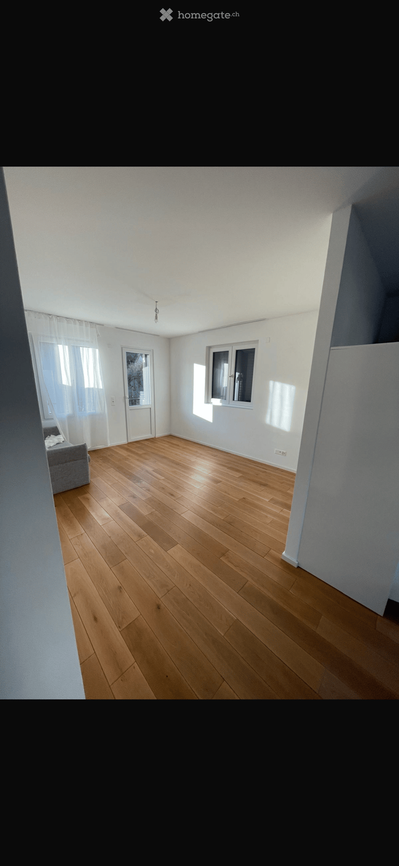 Empty room with wooden flooring, white walls, and large windows with white curtains. No furniture or equipment is present.