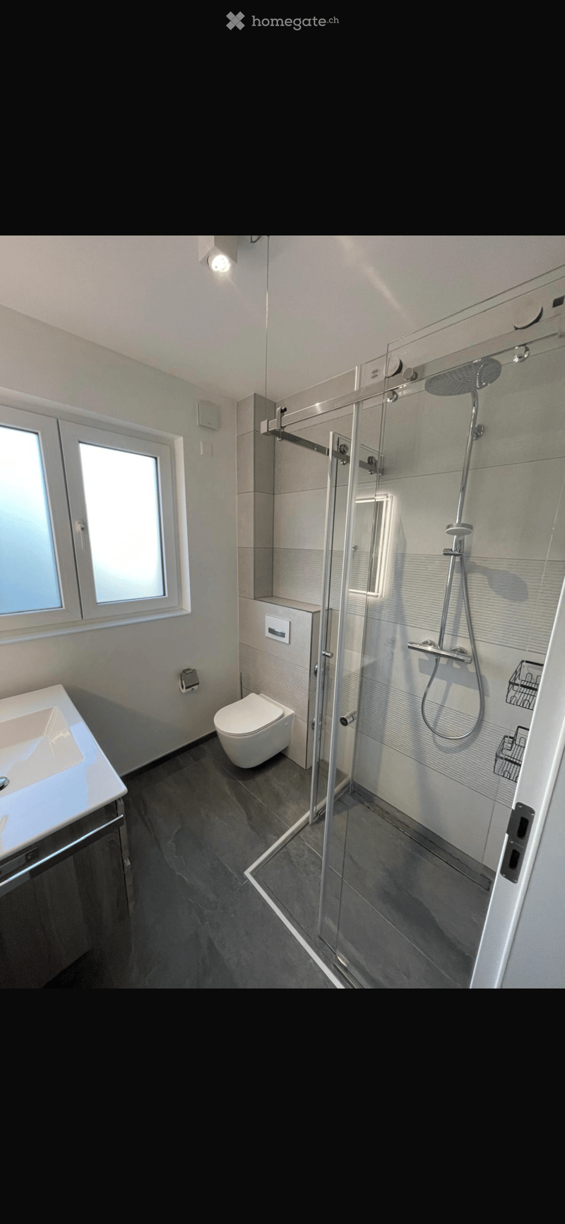 Modern bathroom with a white sink, toilet, and glass-enclosed shower with a handheld showerhead. The space features light-colored tiles and a window for natural light.