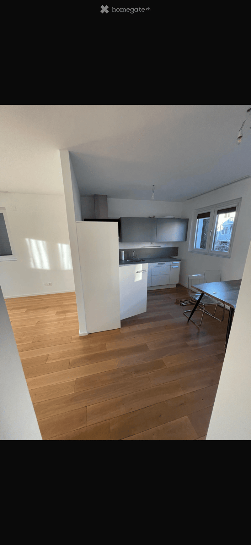 Modern kitchen with white cabinets, a refrigerator, a sink, and a small dining area with a table and chairs. The floor is made of light wooden planks, and the walls are white.