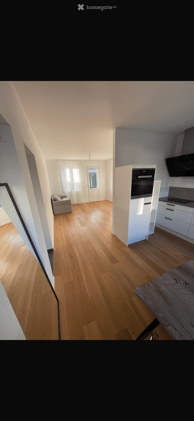A modern, open-plan living and dining area with light wooden flooring, white walls, and large windows. The space includes a sofa, a kitchen area with cabinets and appliances, and a dining table.