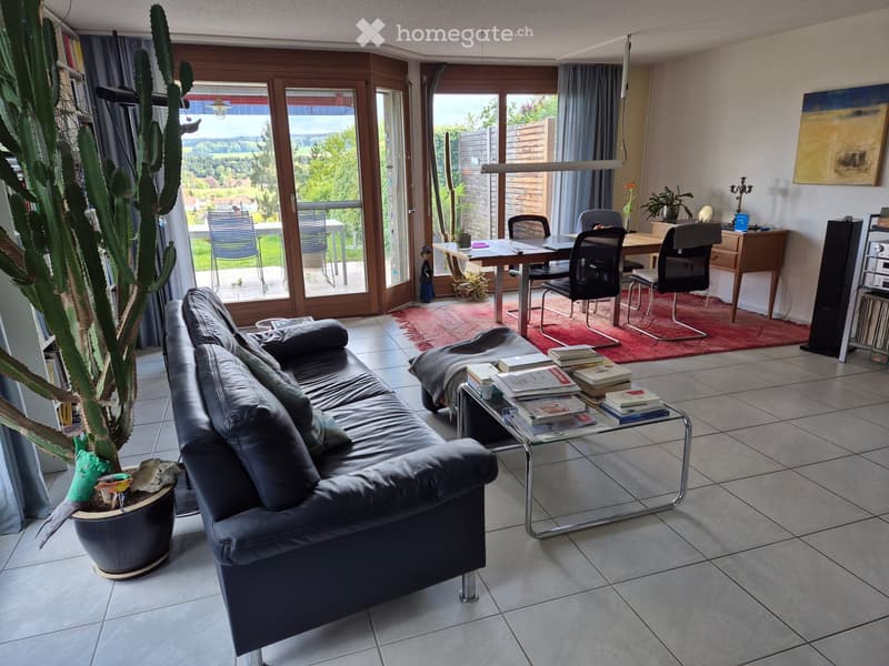 Modern living room with large windows offering a scenic view, featuring a black leather sofa, glass coffee table, dining table with chairs, desk, and a tall cactus plant.