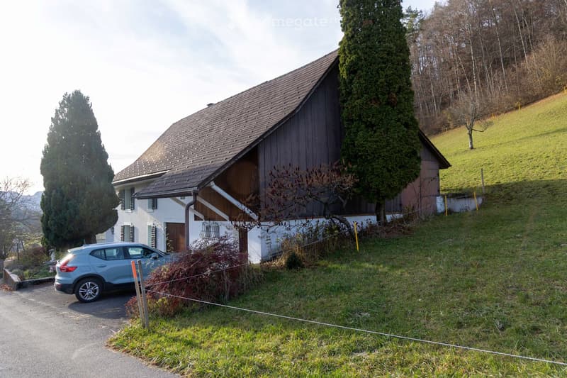 Two-story house with a wooden barn-style extension, surrounded by trees and a grassy slope. A silver SUV is parked in front.