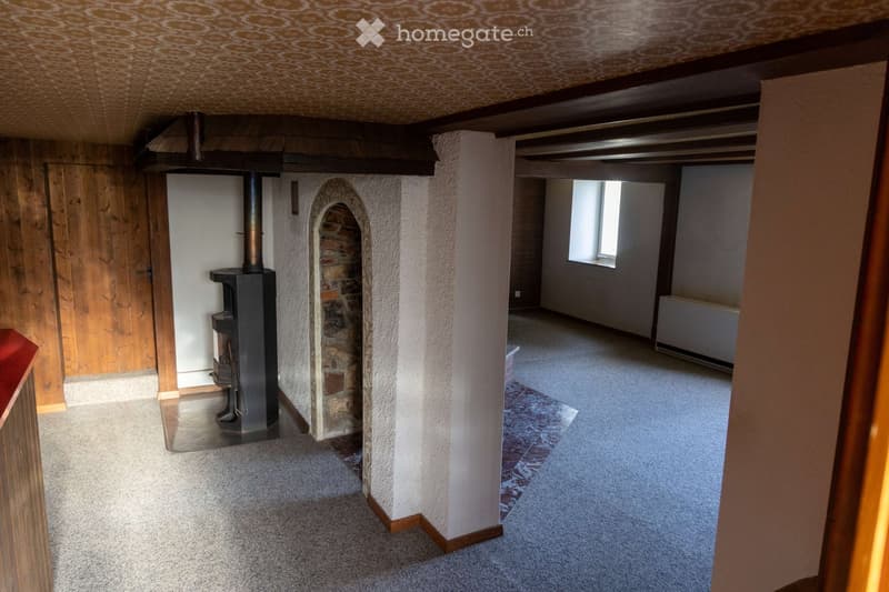 Empty room with carpeted floor, exposed wooden beams, and a fireplace with a chimney. There is a doorway leading to another room with a window and radiator.