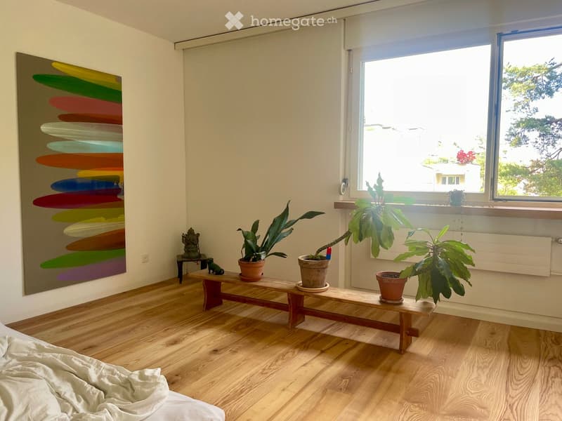 Bright bedroom with wooden floor, large window, bench with potted plants, and colorful abstract painting on wall.