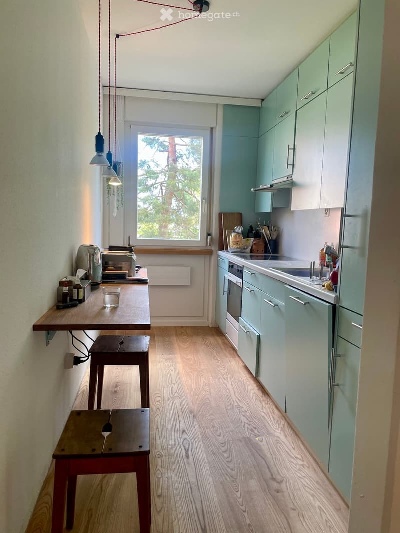 Modern kitchen with light green cabinets, wooden floor, and a small breakfast area with a table and stools. The kitchen features a sink, oven, and various small appliances on the counter.