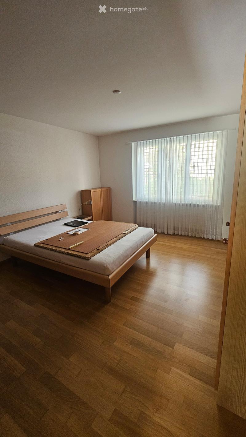 Minimalist bedroom with wooden bed, wardrobe, small table, and lamp, featuring wooden flooring and large window with curtains.
