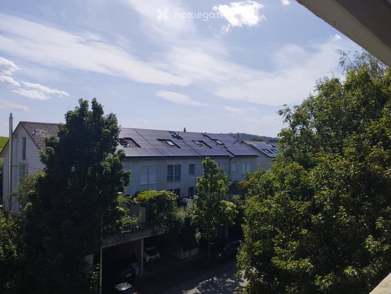 Two-story residential building with solar panels on the roof, surrounded by trees, with a garage and parking area visible.