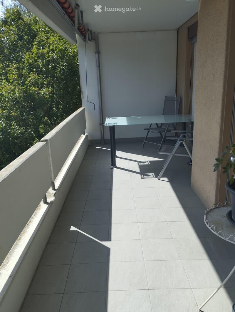 Balcony with tiled floor, glass table, and two folding chairs, overlooking greenery.