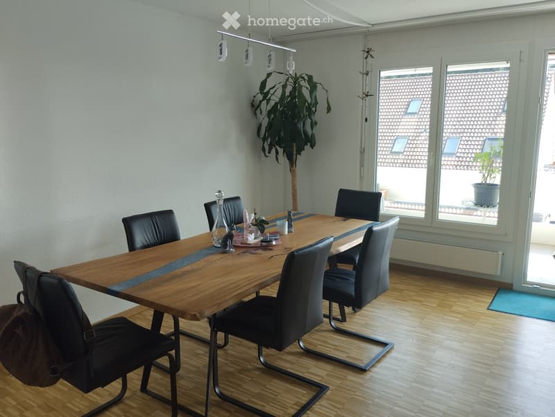 Dining room with a wooden table, black chairs, and a decorative bottle on the table. A large plant is near the window, and the room has wooden flooring and white walls.