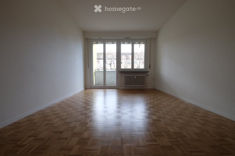 Empty room with wooden parquet flooring, white walls, large window with blinds, and radiator below the window.