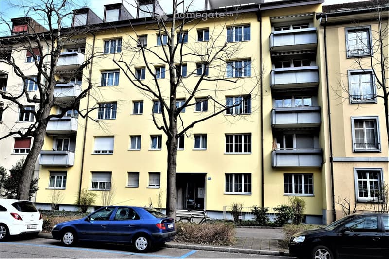 Multi-story residential building with yellow facade, multiple windows, and balconies. A tree is in front of the building, and several cars are parked on the street.