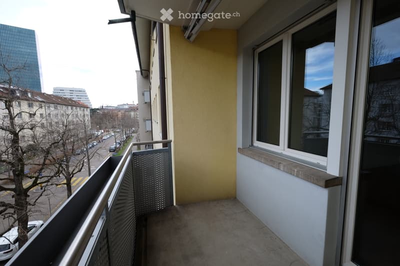 Balcony with metal railing, large windows, overlooking a city street with buildings and parked cars.