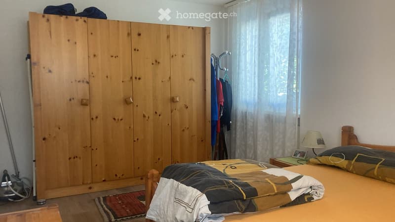 Bedroom with a wooden bed, a large wooden wardrobe, a nightstand with a lamp, and a window with curtains.