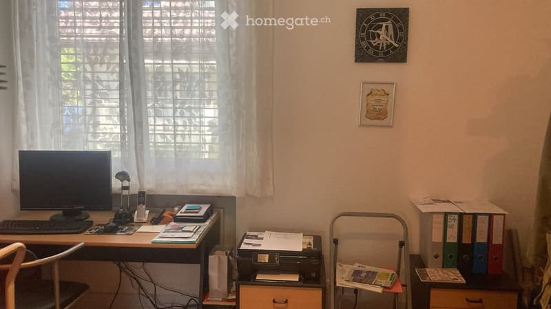 Office space with a desk, computer, printer, filing cabinet, and a clock on the wall. The room has large windows with white curtains.