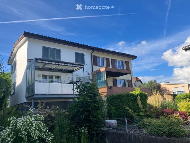 Modern two-story house with white exterior, multiple windows with shutters, and a balcony on the upper level. The house is surrounded by a well-maintained garden with various plants and flowers.
