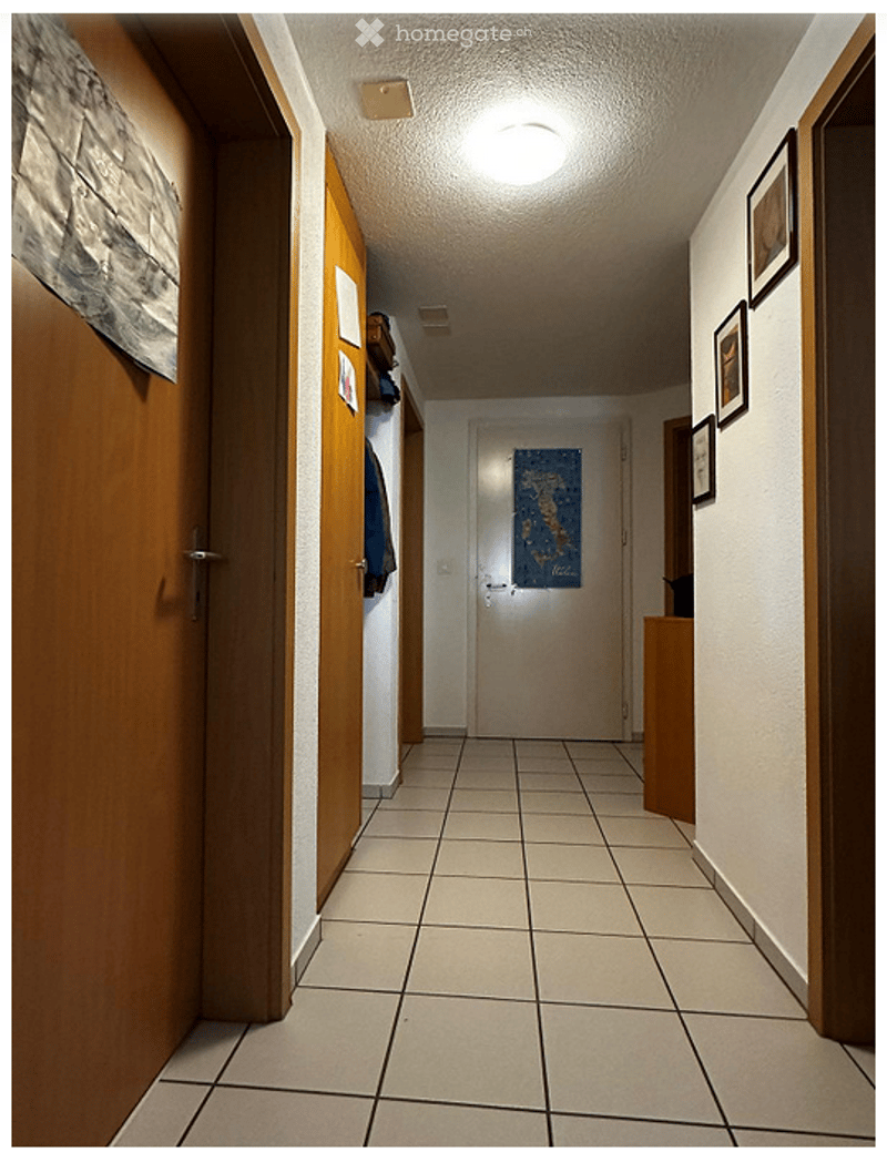 A clean, well-lit hallway with tiled flooring, white walls, and wooden doors on both sides. A small shelf with items is visible on the left, and framed pictures adorn the right wall.