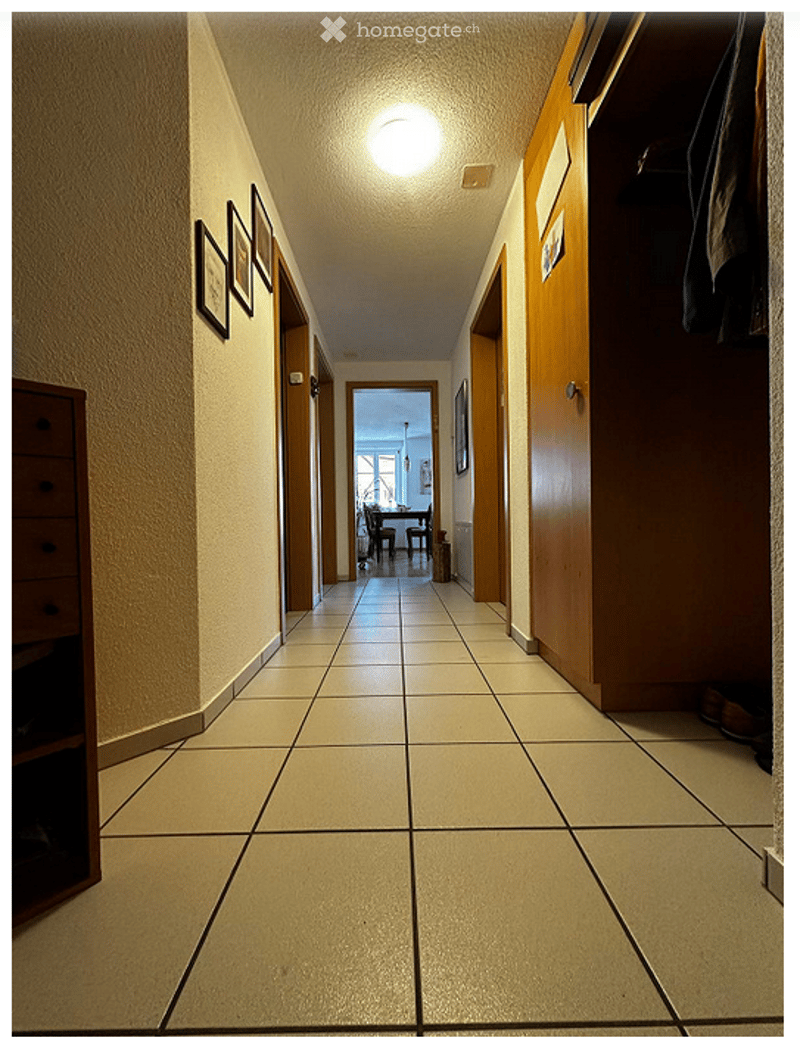A long, tiled hallway with beige walls, framed pictures, and a wooden closet on the right side. The hallway leads to an open area with a dining table and chairs visible in the background.