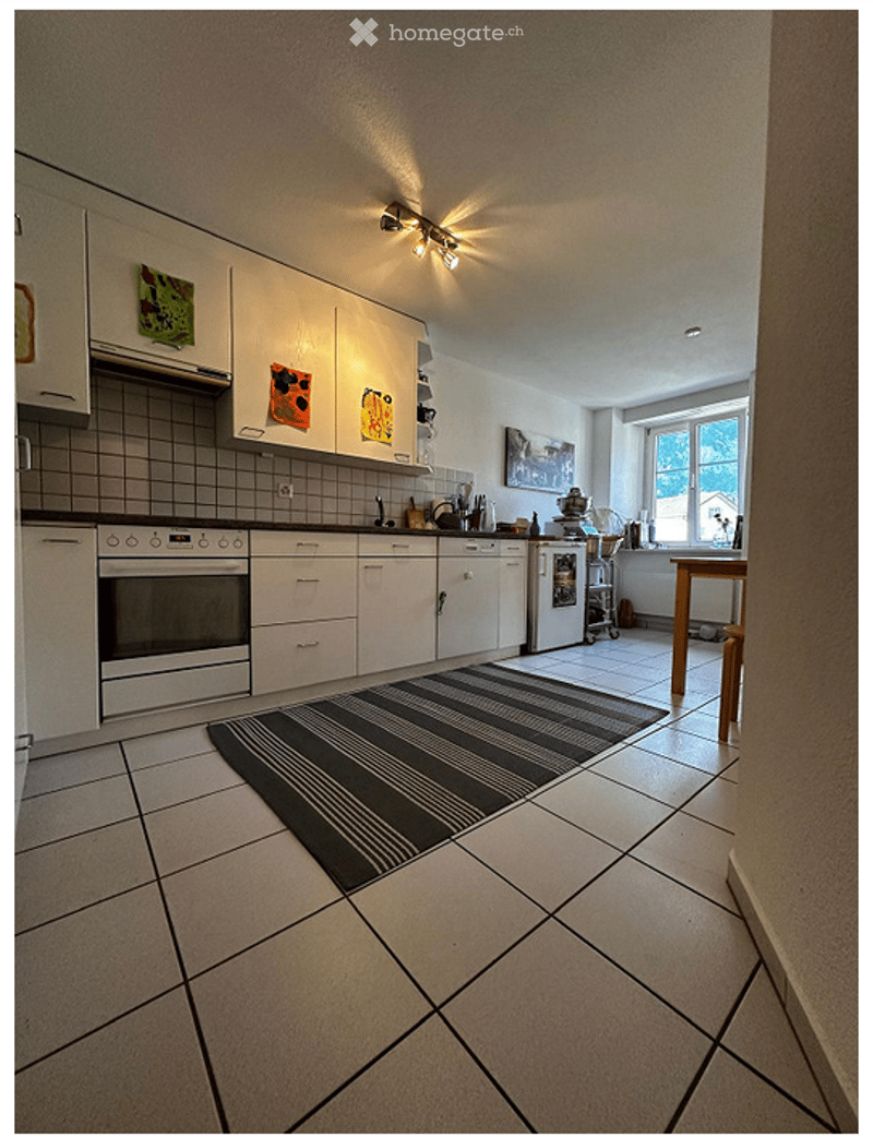 Modern kitchen with white cabinets, tiled walls, and a central island. Features an oven, refrigerator, and a small dining area with a table and chairs. The space is well-lit with recessed lighting and has a black and white striped rug on the floor.