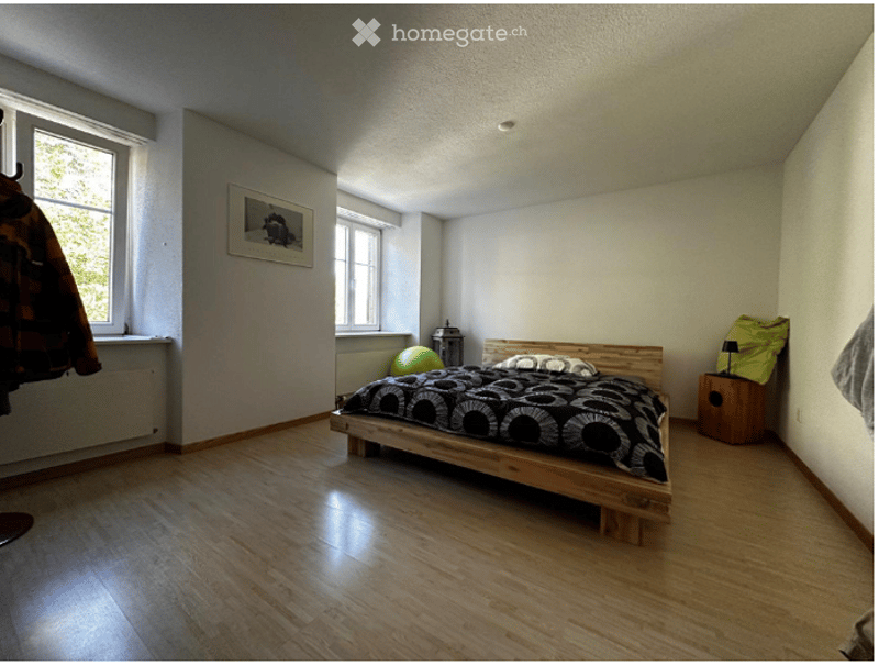 Bright bedroom with wooden flooring, a platform bed with a black and white duvet, a nightstand with a lamp, a picture frame on the wall, and a green exercise ball in the corner.