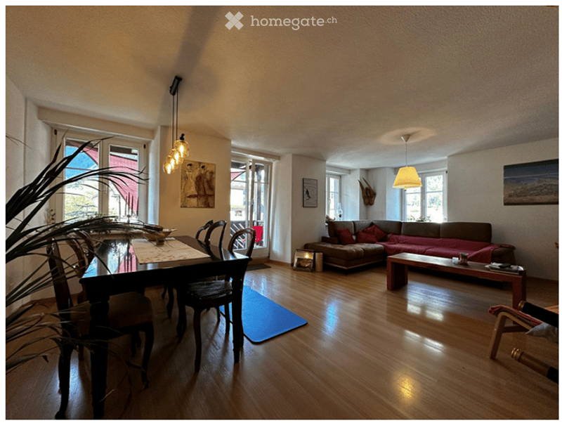 Open-plan living and dining area with wooden flooring, a brown sofa with red cushions, a coffee table, a dining table with chairs, and a floor lamp.