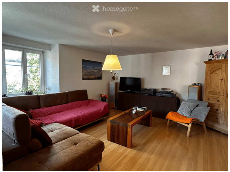 Living room with wooden flooring, a brown sofa with a red blanket, a wooden coffee table, a TV on a stand, a hanging lamp, and a small orange chair.