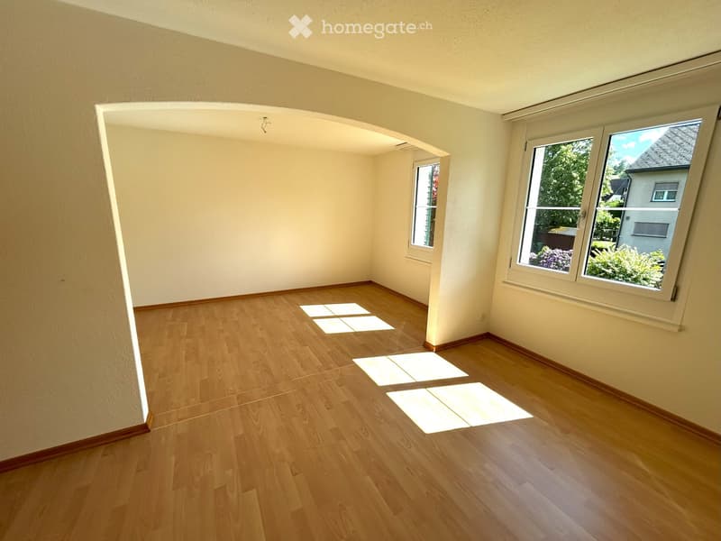 Empty room with wooden flooring and white walls, featuring large windows and an open doorway leading to another space.