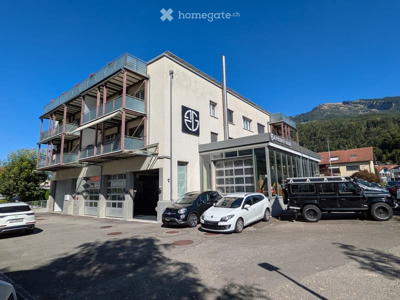 Commercial building with balcony on upper floors, glass doors, logo on the wall, parking lot with multiple cars parked