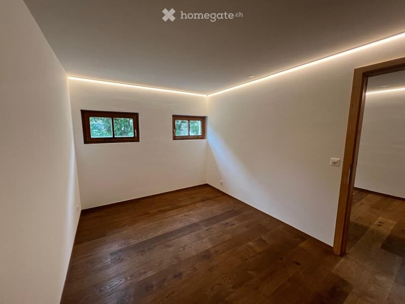 Empty room with wooden flooring, white walls, and recessed lighting. Two small windows with wooden frames are visible, and there is a doorway on the right side.