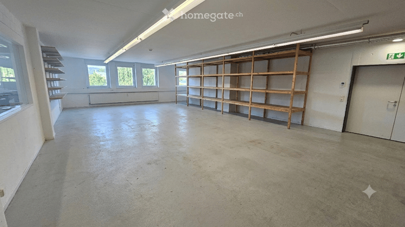 Empty room with concrete floor, white walls, large windows, and wooden shelving units.