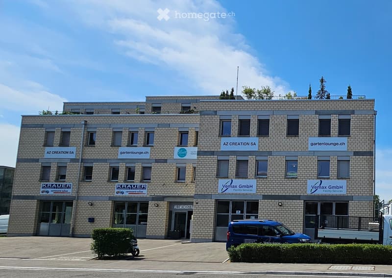 Building. A modern, multi-story commercial building with a brick facade, multiple company logos, and several windows. The building has a flat roof with greenery and is surrounded by a paved area with parked vehicles.