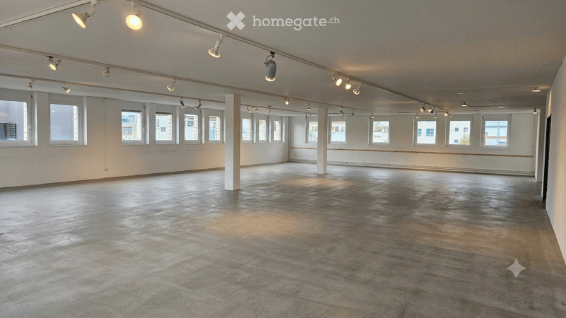 Empty room with concrete floor, white walls, large windows, and track lighting. No furniture or equipment present.