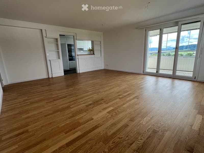 Empty room with wooden flooring, large sliding glass doors leading to a balcony, and a small kitchen area visible in the background.