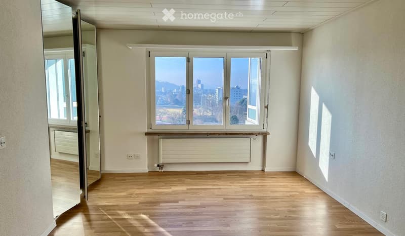 Empty room with wooden flooring, large windows offering a city view, and a radiator under the windows.
