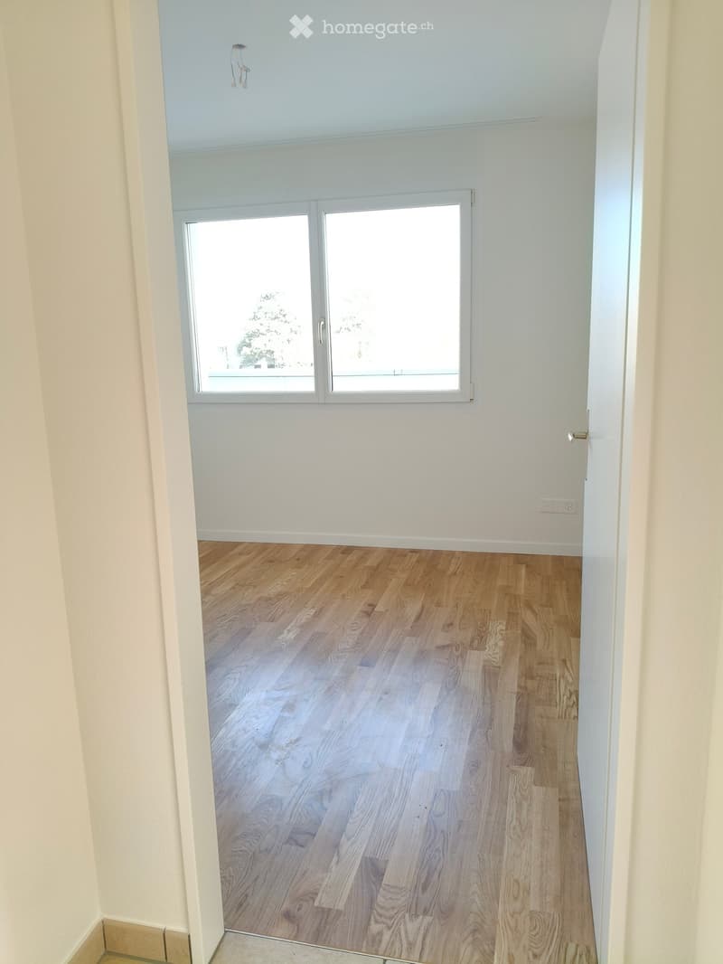 Empty room with light wooden flooring, white walls, and a large window with white frame. No furniture or equipment visible.
