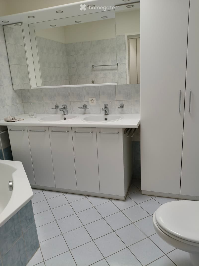 Modern bathroom with white tiled floor, white bathtub, toilet, double sink with mirrored cabinet, and white tiled walls.