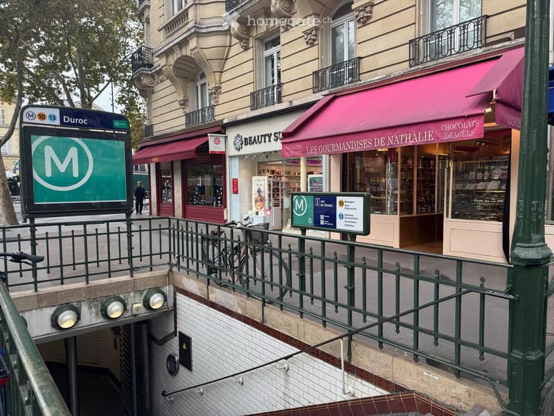 An urban scene with a subway entrance and shops along a street, including a beauty shop and a chocolatier.