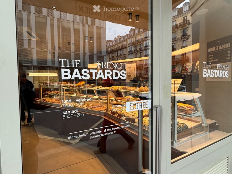 Bakery storefront with glass display cases filled with pastries and bread, signage reading "The French Bastards" and operating hours, located in a city with classic European architecture visible through the windows.