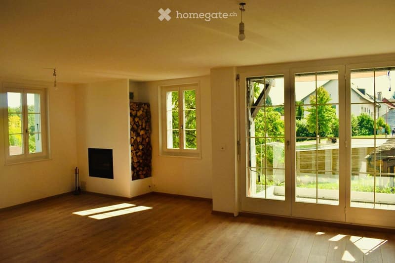 Empty living room with wooden flooring, large windows, and a fireplace. Natural light streams in, and there is a stack of firewood in one corner.