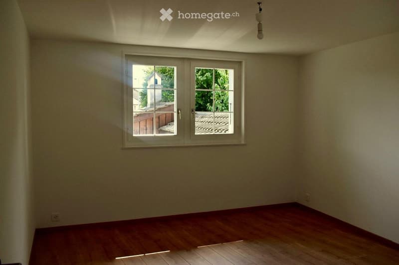 Empty room with wooden flooring and large windows offering a view of a tiled roof and greenery outside.
