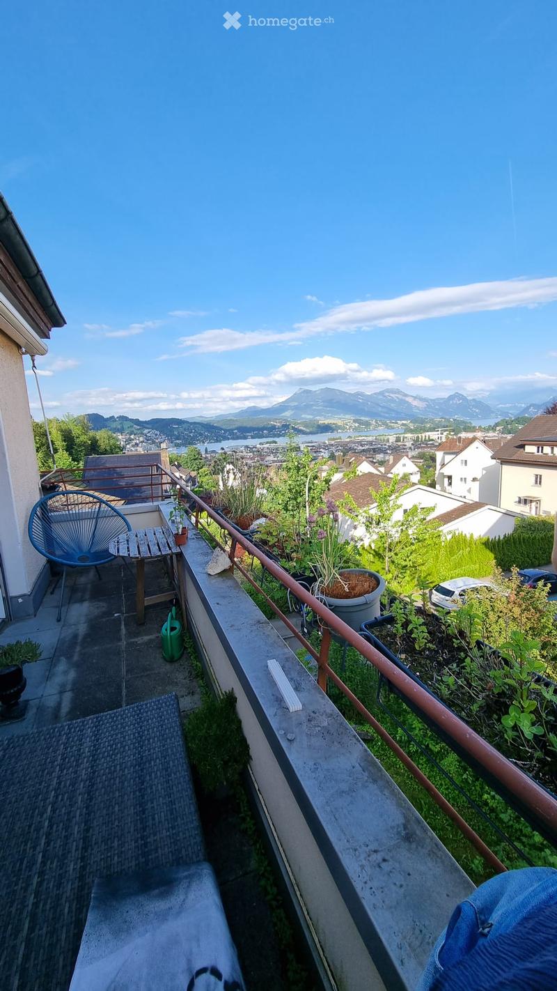 Balcony with a scenic view of mountains and a lake, featuring a blue chair, small wooden table, and potted plants.