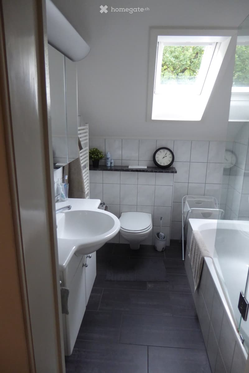 Modern bathroom with white tiles, a sink, toilet, bathtub, and a window letting in natural light. Includes a towel, clock, and small plant on the counter.