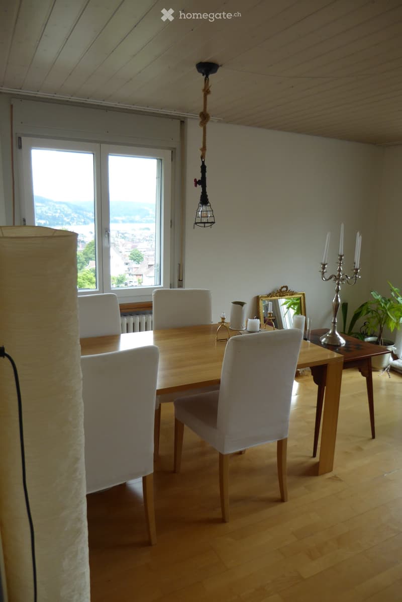 Bright dining area with wooden table, four chairs, view through large windows, decorative items including a candelabra, mirror, and plant.