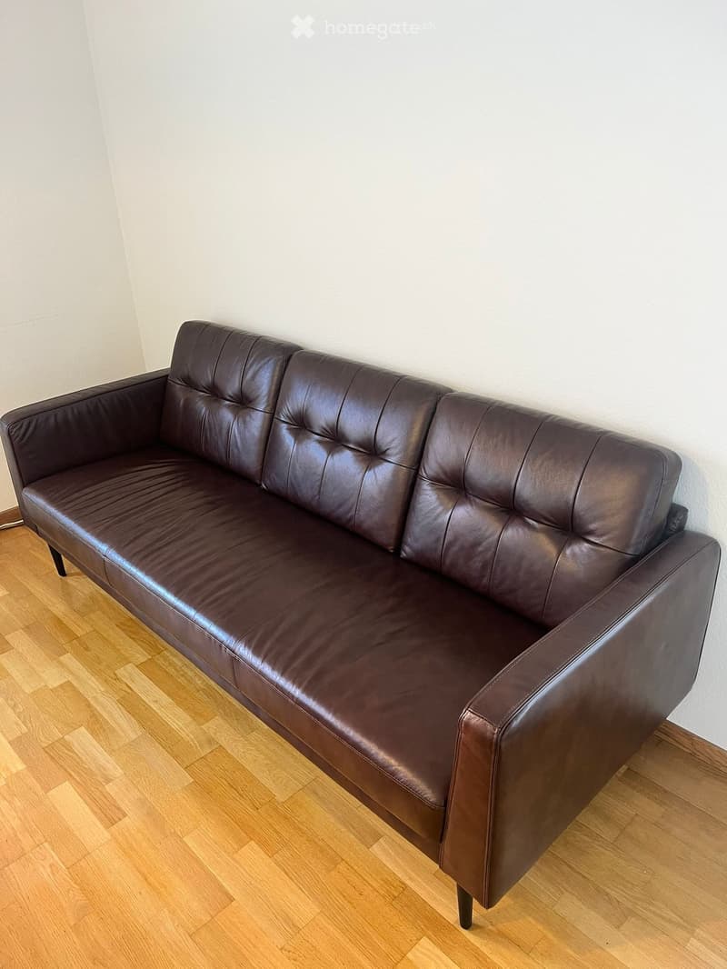 A dark brown leather sofa with tufted backrest and armrests placed on a light wooden floor against a white wall.