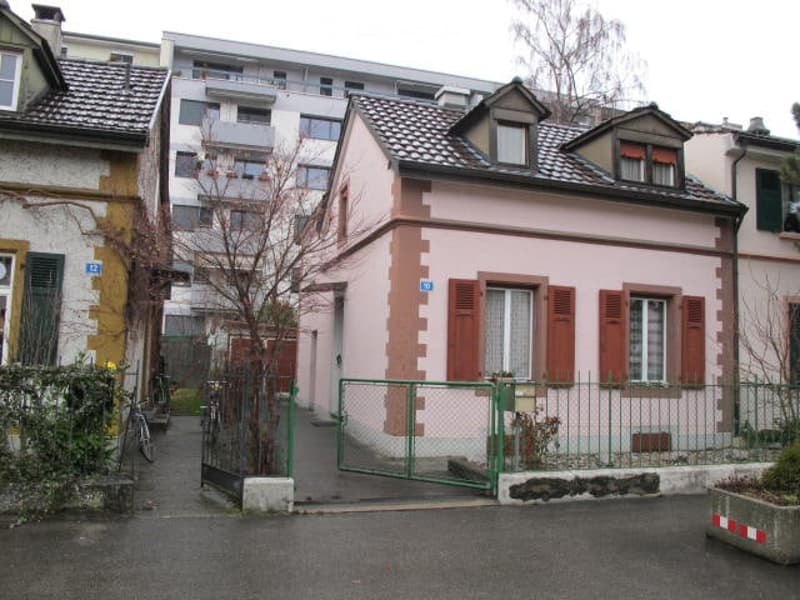 A small, two-story pink house with red shutters and a green gate, situated in a residential area with neighboring buildings and a bicycle parked nearby.