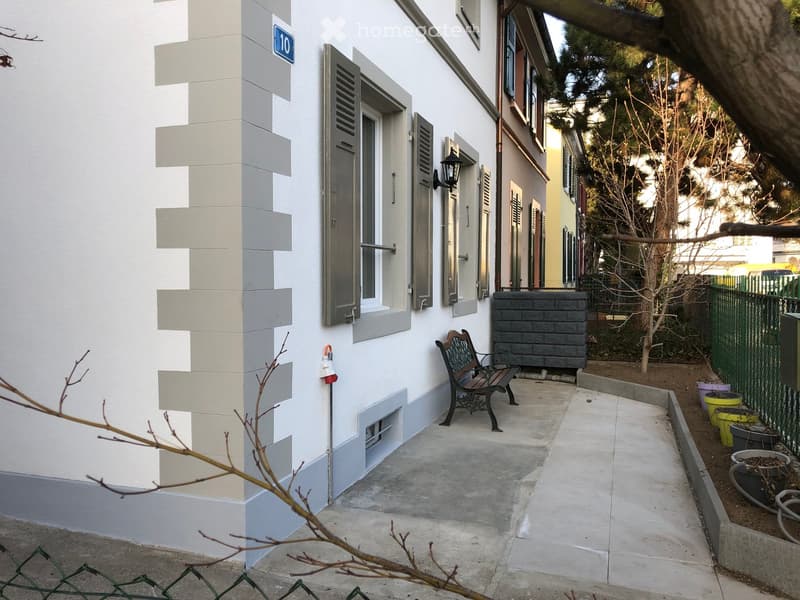 A small outdoor area adjacent to a building, featuring a bench, a grey brick wall, potted plants, and a green fence.
