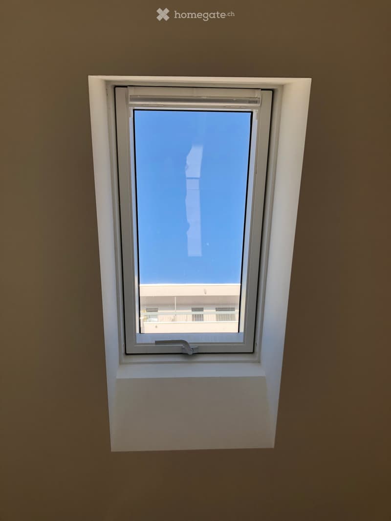 An unfurnished room with a single window showing a view of a building outside. The window is white-framed and has a simple design.
