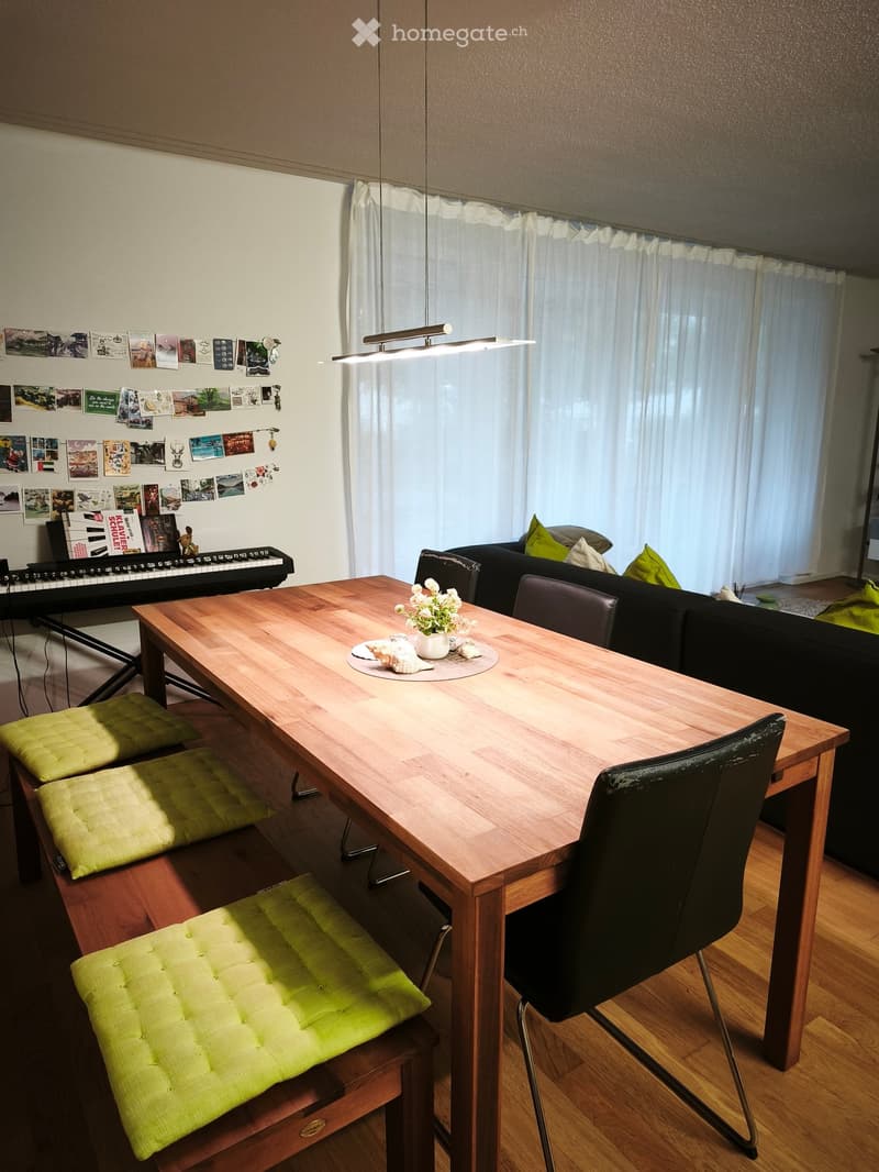 A modern living room with a wooden dining table, black leather chair, green stools, a black sofa, and a small piano. The room features large windows with white curtains and a wall decorated with picture frames and posters.