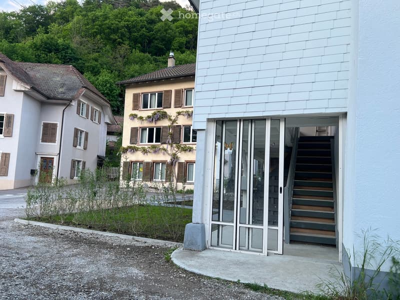 Building. A modern building with a white exterior and a glass door leading to a staircase. The building is surrounded by a small garden with shrubs and a gravel path. In the background, there are two other buildings with brown roofs and wooden shutters. T