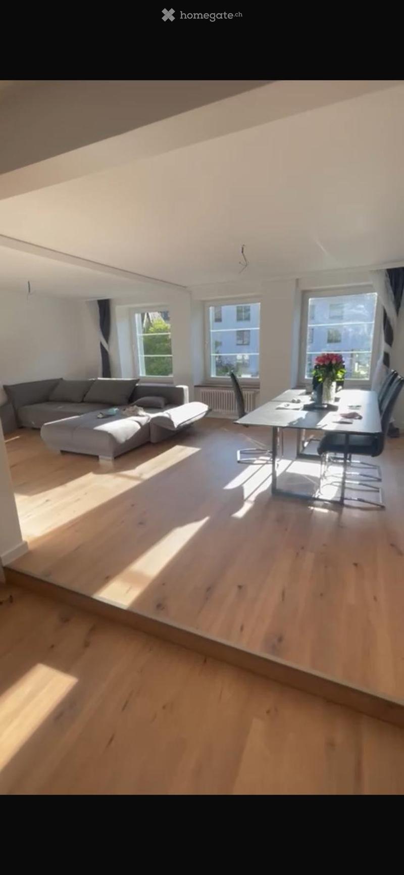 Large, bright living room with wooden floor, windows overlooking outside, sofa, dining table, flowers on table, wooden chairs